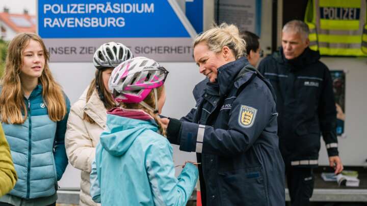 Eine Polizistin schließt einem Kind den Fahrradhelm. Dahinter stehen weitere Polizisten und Kinder. Im Hintergrund steht ein großes Fahrzeug mit der Aufschrift Polizeipräsidium Ravensburg.