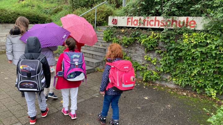 Sechs Kinder laufen mit dem Rücken zur Kamera auf eine Treppe zu. Neben der Treppe hängt ein Schild mit der Aufschrift Flattichschule. Drei der Kinder tragen Schulränzen und zwei Kinder haben einen aufgespannten Regenschirm in der Hand.