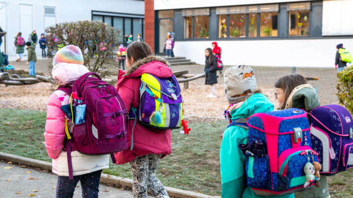 Im Vordergrund laufen vier Kinder mit dem Rücken zur Kamera auf eine Schule zu. Die Kinder tragen Schulränzen.